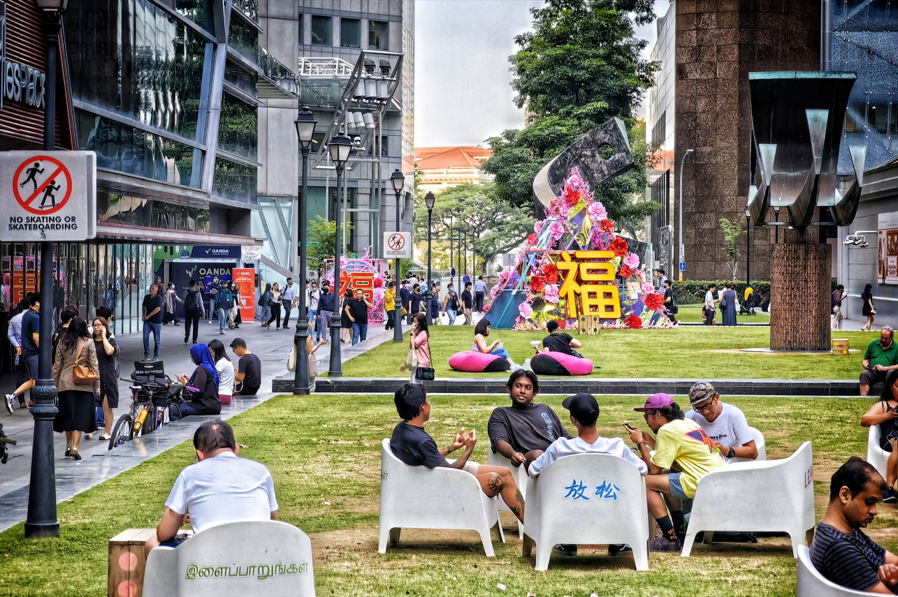 A city park scene with people relaxing on chairs and beanbags in front of a floral display with Chinese characters.